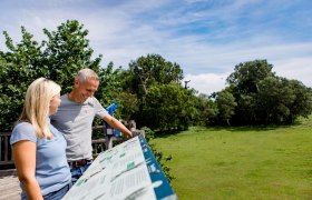 Ausblick Storchenplattform, © Schwarz Koenig Sinzinger Ein Mann und eine Frau stehen auf einer Plattform und betrachten eine Informationstafel. Im Hintergrund sind Bäume und ein blauer Himmel zu sehen.