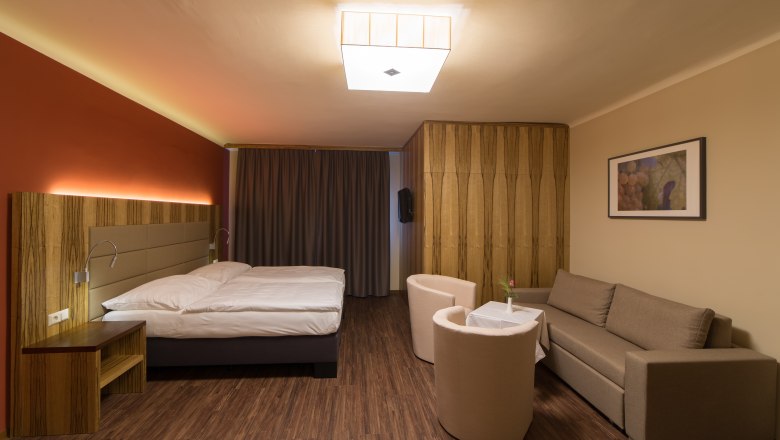 Zimmeransicht, © Fam. Schweinberger Modern eingerichtetes Hotelzimmer mit Doppelbett, Sofa, zwei Sesseln und einem kleinen Tisch. Holzboden und warme Beleuchtung.