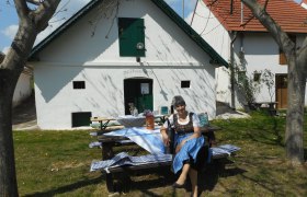 Kellergasse Sierndorf/March, © Franz Rauscher Eine Frau in traditioneller Kleidung sitzt auf einer Bank vor einem weißen Gebäude mit grünen Fensterläden.