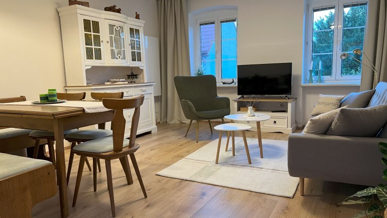 Gemütliches Wohnzimmer mit Holzmöbeln, Sofa, Sessel, Esstisch und Fernseher. Helle Wände und Holzboden.