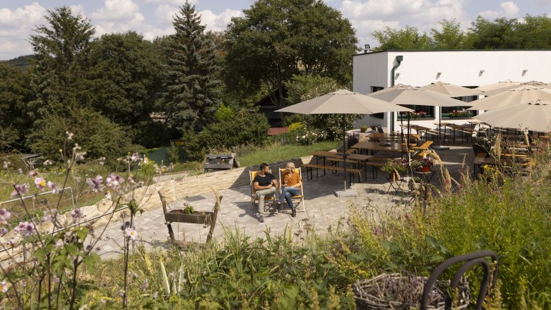 Zwei Personen sitzen auf Liegestühlen in einem Garten mit Sonnenschirmen und Tischen, umgeben von Bäumen und Blumen.