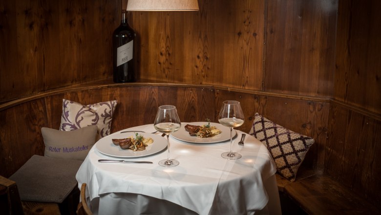 Gemütliche Gaststube mit gedecktem Tisch, zwei Tellern mit Speisen und Weingläsern, Holzwand und Kissen.