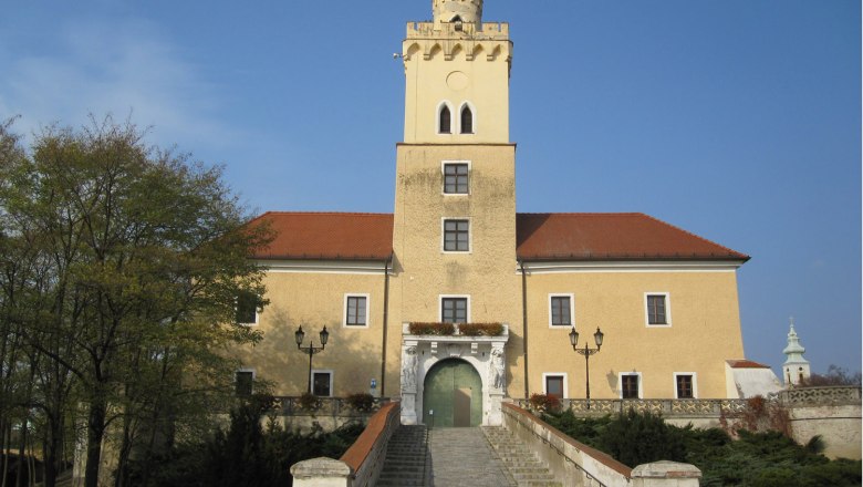 Schloss mit gelber Fassade und rotem Dach, zentraler Turm, breite Steintreppe, blauer Himmel.