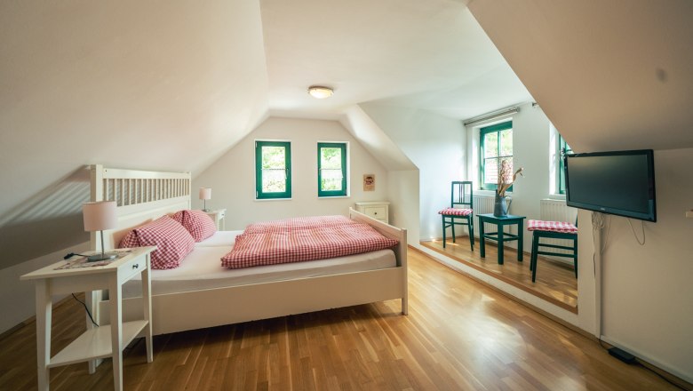 Gem&uuml;tliches Schlafzimmer mit Holzboden, Doppelbett, Nachttischen, zwei St&uuml;hlen und einem Fernseher an der Wand.