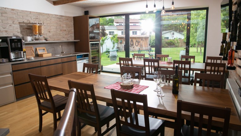 Innenansicht eines modernen Gastraums mit Holztischen und Stühlen, großen Fenstern und Blick auf einen Garten.