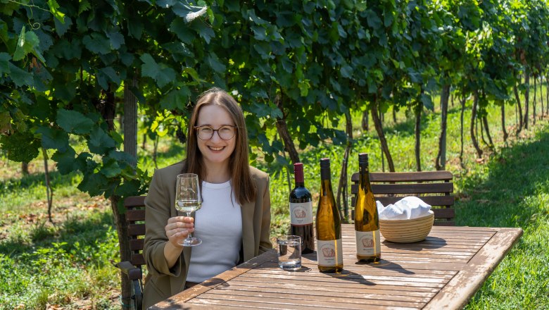 Eine Frau sitzt an einem Holztisch im Weinberg und h&auml;lt ein Weinglas. Auf dem Tisch stehen mehrere Weinflaschen.