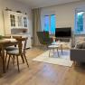 Gemütliches Wohnzimmer mit Holzmöbeln, Sofa, Sessel, Esstisch und Fernseher. Helle Wände und Holzboden.