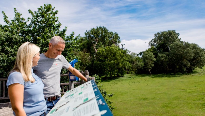 Ein Mann und eine Frau stehen auf einer Plattform und betrachten eine Informationstafel. Im Hintergrund sind B&auml;ume und ein blauer Himmel zu sehen.
