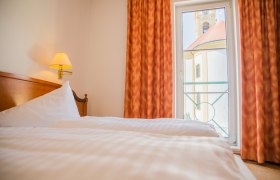 Doppelbett mit weißer Bettwäsche. Im Hintergrund ein Bodentiefes Fenster mit Ausblick auf die Kirche.