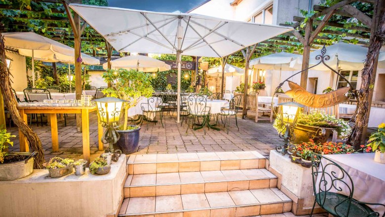 Gemütlicher Gastgarten mit Tischen, Stühlen und Sonnenschirmen, umgeben von Pflanzen und Lichtern.
