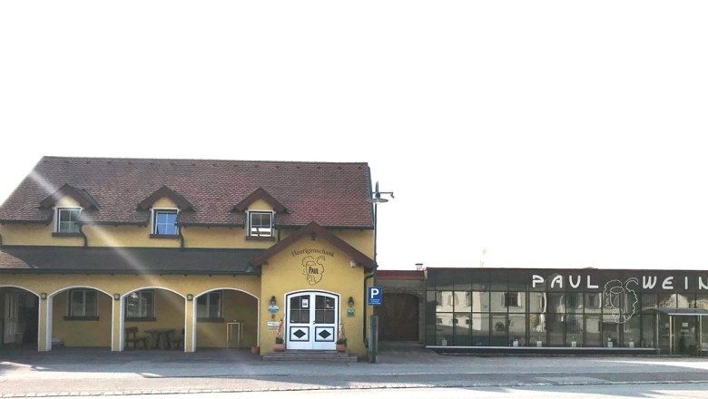 Außenansicht eines gelben Heurigenlokals mit dem Namen 'Paul' und einem modernen Anbau mit der Aufschrift 'Paul Wein'.