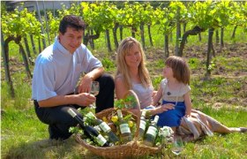 Eine Familie sitzt in einem Weinberg neben einem Korb mit Weinflaschen.