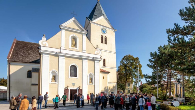 Menschen versammeln sich vor einer Kirche mit Turm und Uhr bei sonnigem Wetter.
