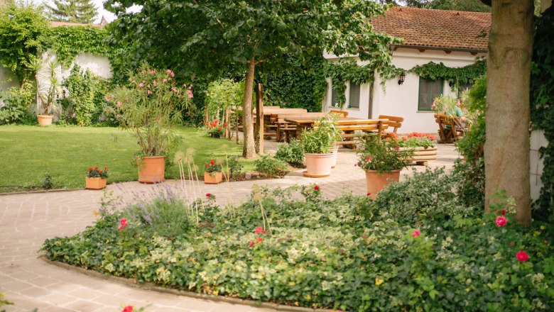 Ein gepflegter Garten mit Rasen, Blumenbeeten und Terrakotta-T&ouml;pfen. Im Hintergrund ein Haus mit Terrasse und Holztischen.