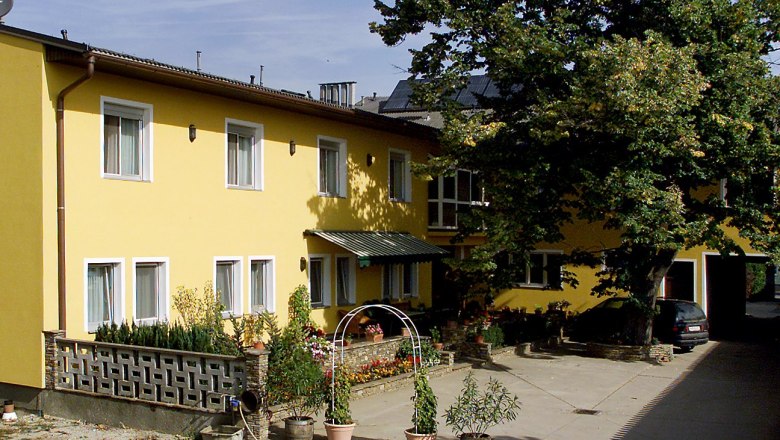 Privatzimmer Familie Holzer, © Fam. Holzer Gelbes Gebäude mit mehreren Fenstern, Pflanzen im Vordergrund und einem großen Baum daneben.