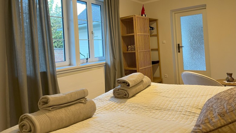 Ein gem&uuml;tliches Schlafzimmer mit einem Bett, auf dem Handt&uuml;cher liegen, einem Fenster mit Vorh&auml;ngen und einem Regal mit Dekoration.