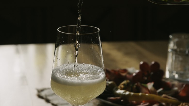 Ein Glas Wei&szlig;wein wird auf einem Tisch eingeschenkt, im Hintergrund ein Teller mit Snacks.
