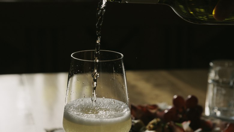 Ein Glas Weißwein wird auf einem Tisch eingeschenkt, im Hintergrund ein Teller mit Snacks.