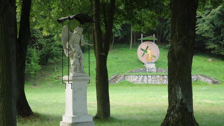 Statue eines Engels mit Kind im Vordergrund, im Hintergrund eine weitere Statue auf einem Podest in einem bewaldeten Park.