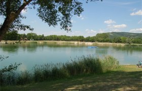 Ein ruhiger See mit Schilf im Vordergrund und bewaldeten Hügeln im Hintergrund.