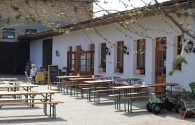 Innenhof einer Buschenschank mit leeren Holzbänken und einem Tisch, zwei Personen sitzen an einem Tisch, ein Spielzeugtraktor steht daneben.