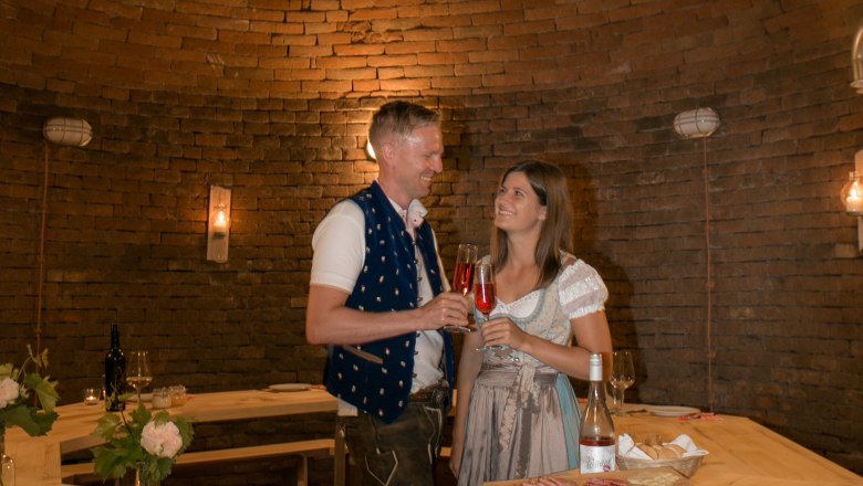 Ein Paar in traditioneller Kleidung stößt mit Getränken in einem rustikalen, gewölbten Raum an.