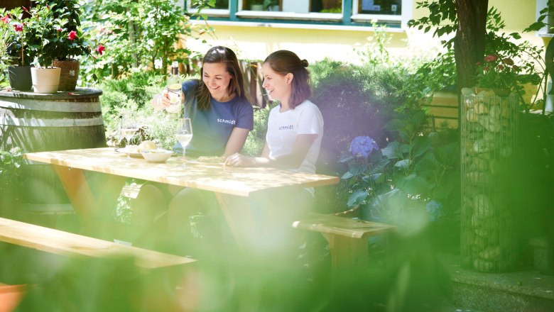 Zwei Frauen sitzen an einem Holztisch im Garten und genie&szlig;en Wein.