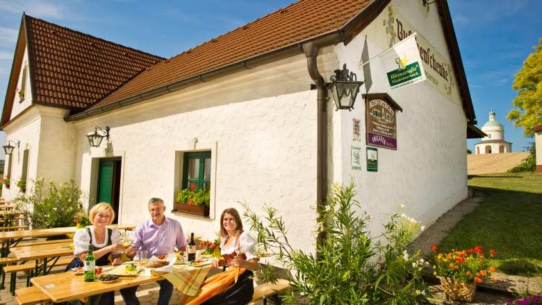 Drei Personen sitzen an einem Tisch vor einem Weingut und genießen eine Mahlzeit im Freien.