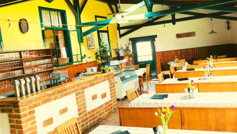 Innenansicht eines gemütlichen Gastraums mit Holztischen, gelben Wänden und einer Bar aus Backstein.