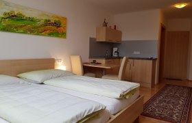 Ein modernes Zimmer mit zwei Betten, einem Schreibtisch, einer kleinen Küchenzeile und einem bunten Gemälde an der Wand.