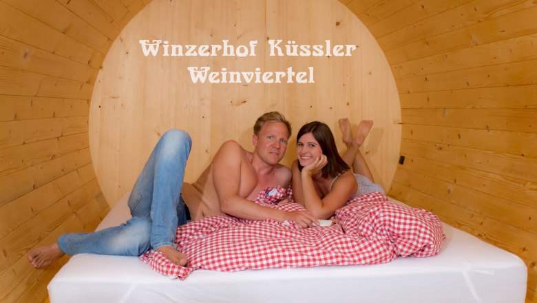 Ein Paar liegt in einem großen Holzfassbett mit rot-weiß kariertem Bettzeug. Im Hintergrund steht 'Winzerhof Küssler Weinviertel'.