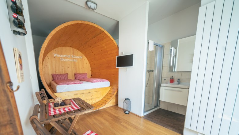 Innenansicht einer Suite mit Bett in einem gro&szlig;en Weinfass, kleinem Tisch mit St&uuml;hlen und Badezimmer.