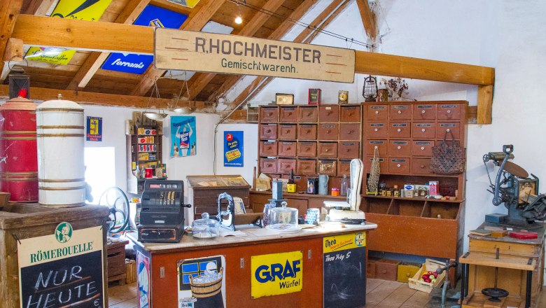 Innenansicht eines alten Gemischtwarenladens mit Holzm&ouml;beln, einer Registrierkasse und Werbeschildern an den W&auml;nden.