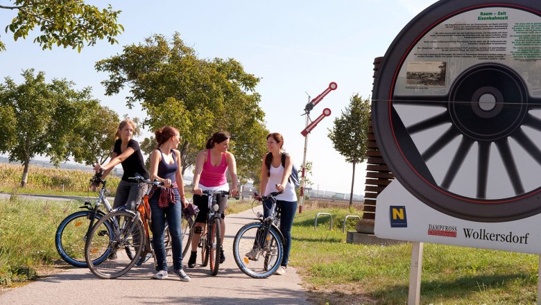 Vier Frauen mit Fahrrädern auf einem Radweg neben einem großen Rad-Denkmal in Wolkersdorf.