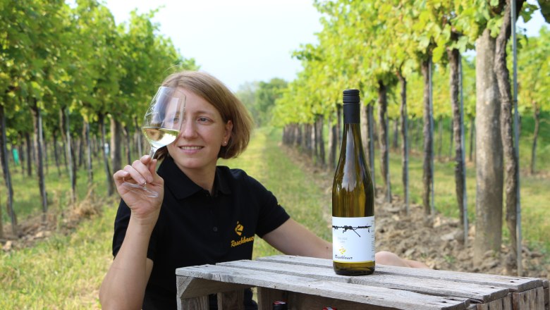 Eine Frau im Weinberg hält ein Glas Weißwein und sitzt neben einer Weinflasche auf einer Holzkiste.
