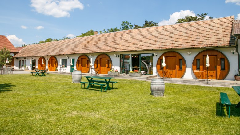 Ein langes Geb&auml;ude mit runden Holzt&uuml;ren und einem gepflegten Rasen davor, auf dem Picknicktische und Weinf&auml;sser stehen.