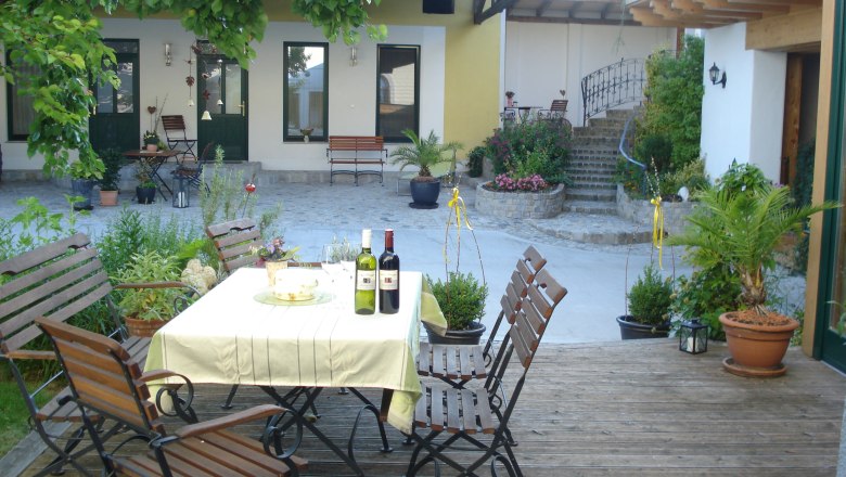 Ein gemütlicher Innenhof mit Tisch, Stühlen und Weinflaschen auf einem Holzboden, umgeben von Pflanzen und einem Gebäude im Hintergrund.