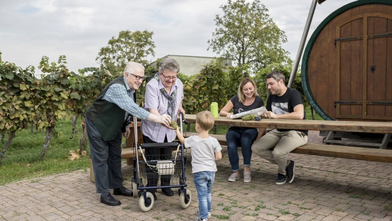 Eine Familie sitzt an einem Holztisch im Freien, umgeben von Weinreben. Ein älteres Paar gibt einem Kind Trauben.