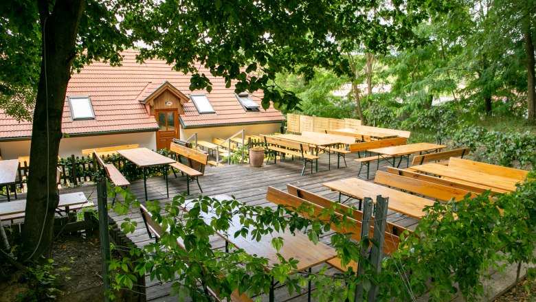 Ein leerer Biergarten mit Holzbänken und Tischen, umgeben von Bäumen und einem Gebäude mit rotem Dach im Hintergrund.