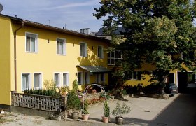 Gelbes Gebäude mit mehreren Fenstern, Pflanzen im Vordergrund und einem großen Baum daneben.