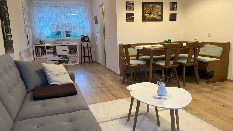 Gem&uuml;tliches Wohnzimmer mit grauem Sofa, Esstisch und St&uuml;hlen, B&uuml;cherregal und Fenster mit Gardinen.