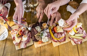 Mehrere Hände greifen nach Aufschnitt und Käse auf Holzplatten.