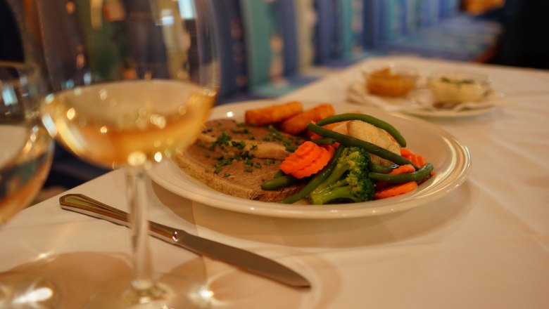 Ein Teller mit Fleisch, Gem&uuml;se und Kartoffeln auf einem Tisch, daneben ein Glas Wei&szlig;wein.