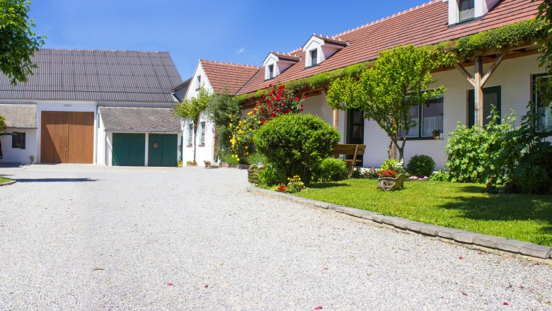 Ein idyllisches Weingut mit Kiesweg, bl&uuml;henden G&auml;rten und traditionellen Geb&auml;uden unter blauem Himmel.