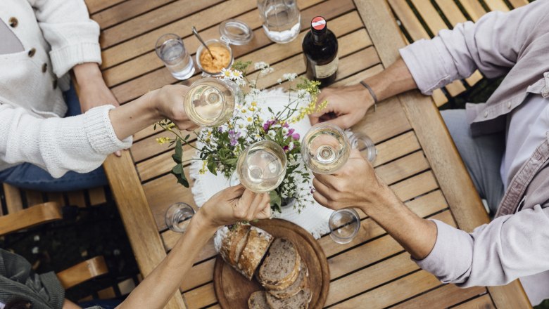 Drei Personen sto&szlig;en mit Weingl&auml;sern an einem Holztisch an, auf dem Brot, Wasser und Blumen stehen.