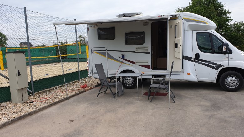 Wohnmobil mit ausgefahrener Markise und Campingm&ouml;beln auf einem Stellplatz neben einem Volleyballfeld.