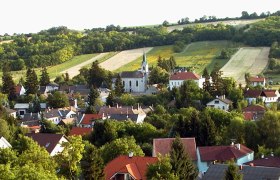 Panorama von Unterolberndorf mit Kirche und umliegenden H&auml;usern vor gr&uuml;nen H&uuml;geln.