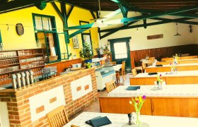 Innenansicht eines gemütlichen Gastraums mit Holztischen, gelben Wänden und einer Bar aus Backstein.