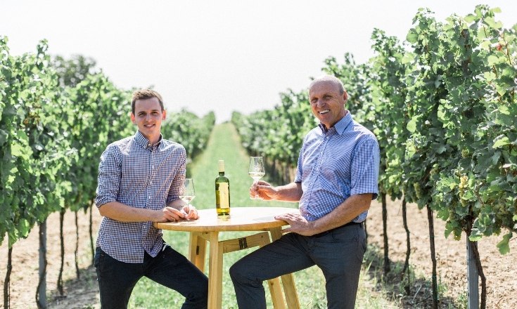 Zwei Männer sitzen an einem Tisch im Weinberg mit Weingläsern.