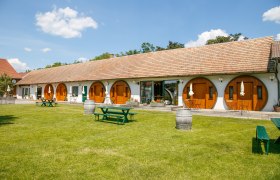 Ein langes Geb&auml;ude mit runden Holzt&uuml;ren und einem gepflegten Rasen davor, auf dem Picknicktische und Weinf&auml;sser stehen.